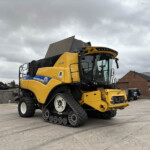 Large yellow New Holland combine harvester with crawler tracks parked in an industrial yard under a cloudy sky, showing its cab and front wheels.