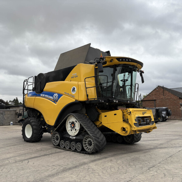 Large yellow New Holland combine harvester with crawler tracks parked in an industrial yard under a cloudy sky, showing its cab and front wheels.