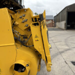 Close-up of yellow industrial machine with hydraulic hoses and bolts; an open workshop building is in the background.