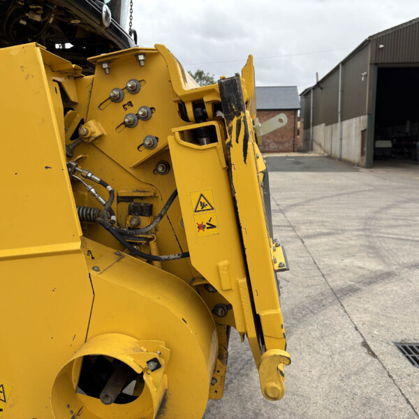 Close-up of yellow industrial machine with hydraulic hoses and bolts; an open workshop building is in the background.