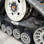 Close-up of a tracked construction machine's wheel and track system, showing bolts and a yellow body behind it.