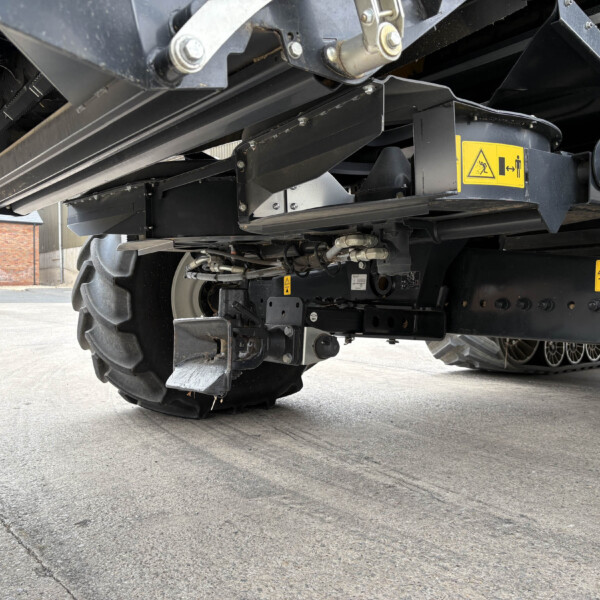 Underside of a large industrial machine with black metal beams and warning labels, tire visible in background behind the equipment.