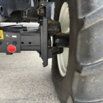 Close-up of a heavy-machine hitch area with a red emergency stop button, yellow warning label, and dirt-caked metal components in the foreground.