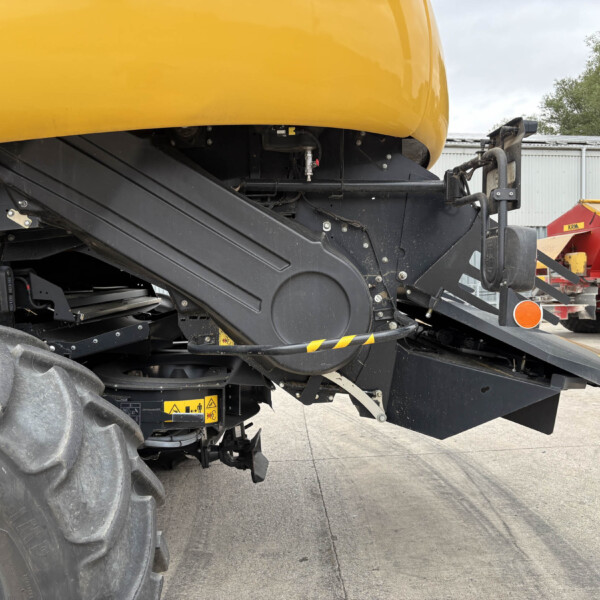 Close-up of the orange agricultural machine’s underside showing black chassis, hoses, and attachment arms, with a red trailer in the background.