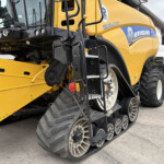 Close-up side view of a yellow New Holland combine harvester with tracked wheels and access ladder, showing branding and machinery details on a farm yard.