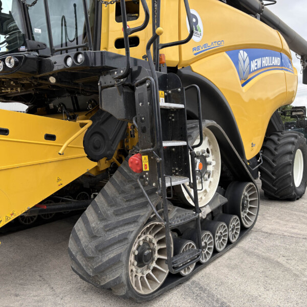 Close-up side view of a yellow New Holland combine harvester with tracked wheels and access ladder, showing branding and machinery details on a farm yard.