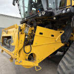 Close-up of a yellow construction machine with a black cab, hydraulic hoses, and warning decals, parked beside a gray building.