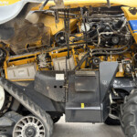 Close-up of the exposed belts, pulleys, and hydraulic lines inside a yellow agricultural combine harvester engine compartment.