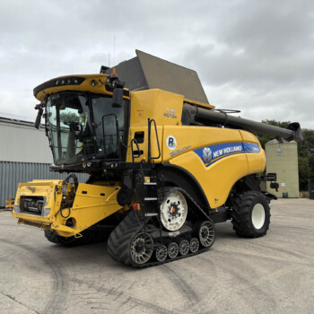 Front three-quarter view of a large yellow New Holland combine harvester with its header attached, parked in an industrial yard on a cloudy day.