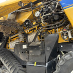 Close-up view of an exposed belt drive and hydraulic lines inside a yellow agricultural machine, showing pulleys, hoses, and metal components.