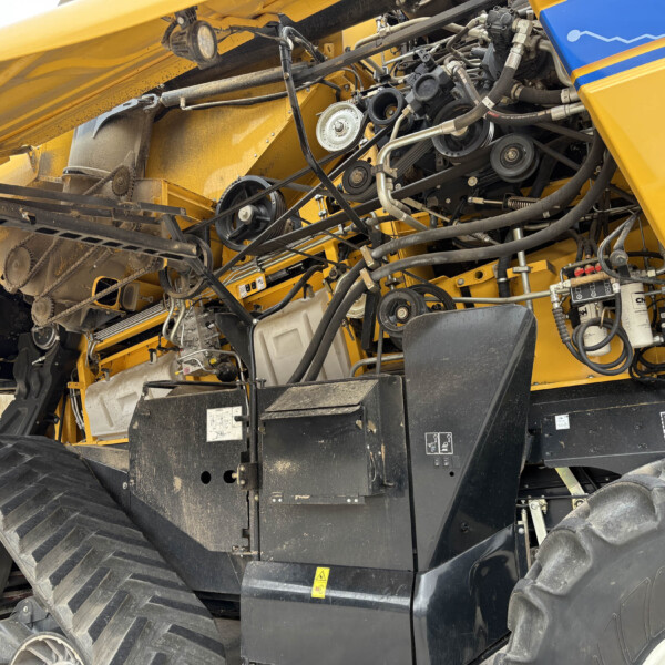 Close-up view of an exposed belt drive and hydraulic lines inside a yellow agricultural machine, showing pulleys, hoses, and metal components.