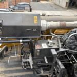 Close-up of heavy machinery hydraulic components and hoses with a yellow body in an industrial yard setting behind parked vehicles.