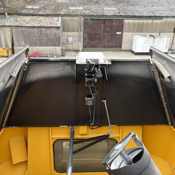 Top-down view of a yellow industrial hopper/conveyor system opened to reveal a dark interior, surrounded by concrete walls and corrugated metal buildings. Industrial yard scene with a large yellow machine, black hopper, and visible pipes and controls between warehouse walls. Overhead perspective of factory equipment: a yellow component with a large black hopper, located amid snow-dusted roofed sheds. View from the operator area into a large yellow hopper with mechanical arms and cables, set in an industrial courtyard. Wide shot of an industrial site featuring yellow machinery and a dark open hopper, with corrugated metal sheds and a white generator in the background.