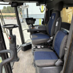 Interior of a construction machine cab with blue upholstered seats, steering wheel, and control panel visible through the windshield