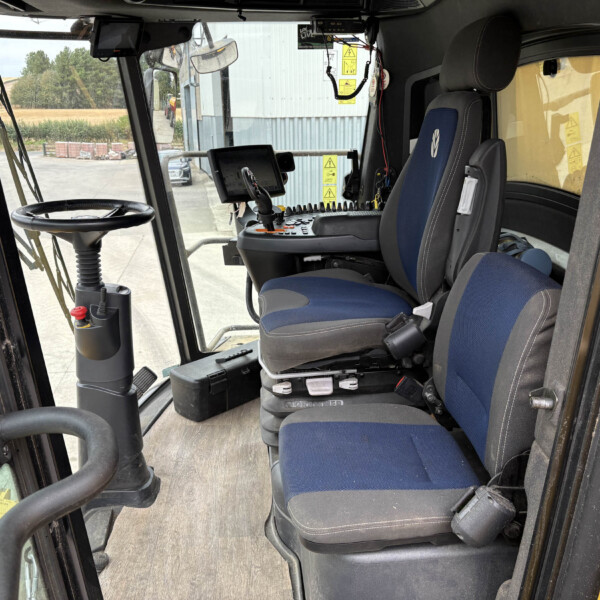 Interior of a construction machine cab with blue upholstered seats, steering wheel, and control panel visible through the windshield