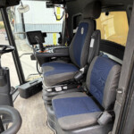 Inside the cabin of a construction vehicle showing a blue-and-gray seat, joystick control, display screen, and nearby control panel by the window.