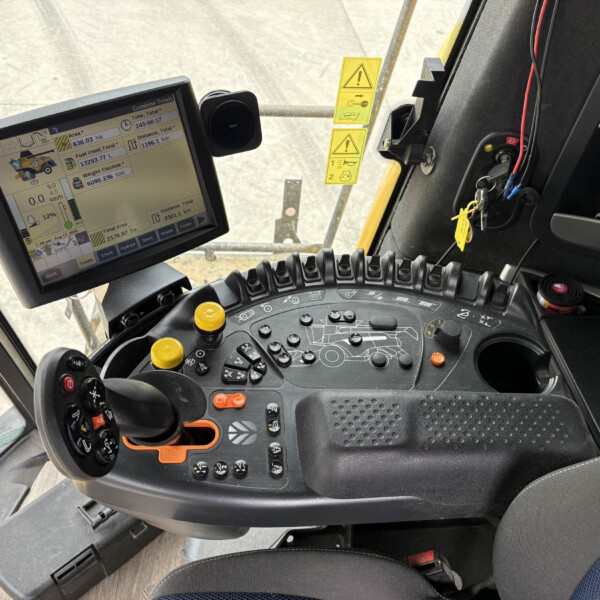 Inside a tractor cab with a mounted touchscreen display, levers, keys, and yellow safety stickers near the windshield.