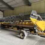 Yellow agricultural header on a trailer inside a large warehouse or workshop with metal walls and ceiling beams.