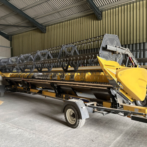 Yellow agricultural header on a trailer inside a large warehouse or workshop with metal walls and ceiling beams.