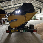 Rear view of a yellow New Holland combine harvester inside a large shed, with a green tractor in the background.