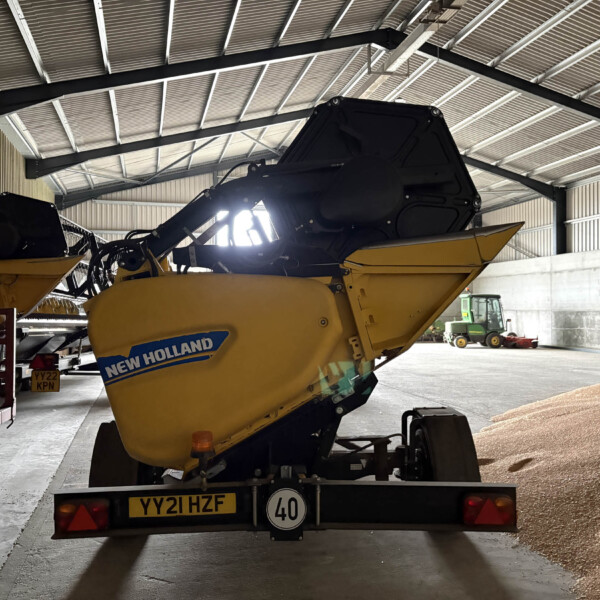 Yellow New Holland combine harvester mounted on a trailer inside a large industrial shed, with gravel on the floor and a green tractor visible in the background on the right.