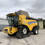 Yellow New Holland combine harvester with tracked wheels parked in an industrial yard, front header attached, under a cloudy sky.