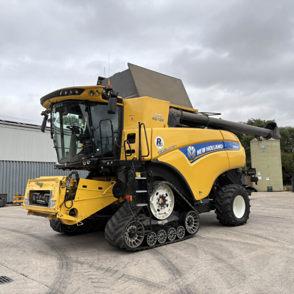 Yellow New Holland combine harvester with tracked wheels parked in an industrial yard, front header attached, under a cloudy sky.