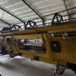 Yellow agricultural combine header mounted on a trailer inside a workshop, showing blade guards and conveyor components.