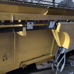 Close-up of a yellow industrial machine showing metal brackets, bolts, and hanging cables in a factory setting.