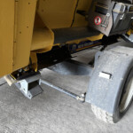 Close-up of a yellow construction/industrial machine showing a black belt-like component, metal steps, and a wheel with the New Holland branding visible on the frame.