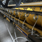 Close-up of a New Holland combine header with metal finger guards and rotating tines on a yellow machine in a workshop setting