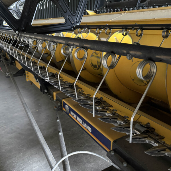Close-up of a New Holland combine header with metal finger guards and rotating tines on a yellow machine in a workshop setting