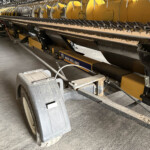 Close-up of a yellow New Holland header underside with conveyor chains and steel support frame in a workshop setting.