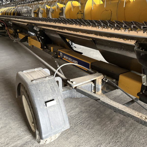 Close-up of a yellow New Holland header underside with conveyor chains and steel support frame in a workshop setting.