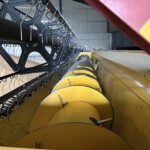 Close-up of a combine harvester header with curved metal tines and yellow guards in a shed-like space