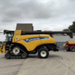 Yellow New Holland combine harvester parked in a equipment yard under a cloudy sky, side view showing tracks and header.