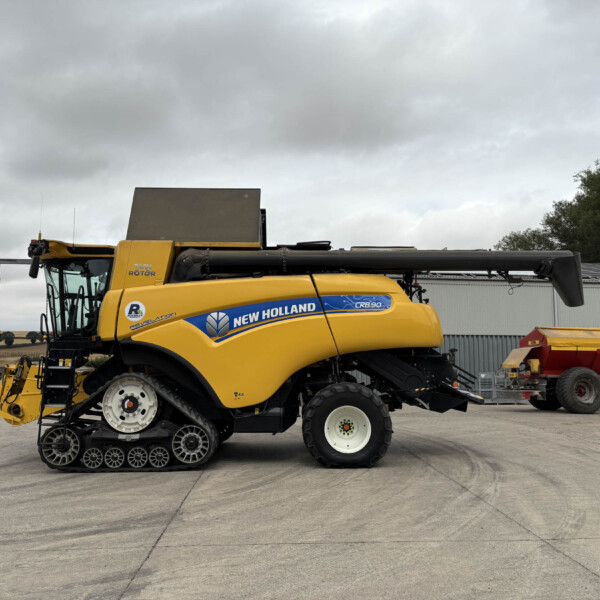 Yellow New Holland combine harvester parked in a equipment yard under a cloudy sky, side view showing tracks and header.