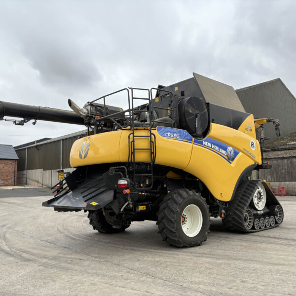Yellow New Holland combine harvester with an extended unloading auger parked in a yard beside warehouses and brick buildings.