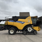 Yellow New Holland CR8.90 combine harvester with crawler tracks parked in an industrial yard against a cloudy sky, side view showing branding and machinery.