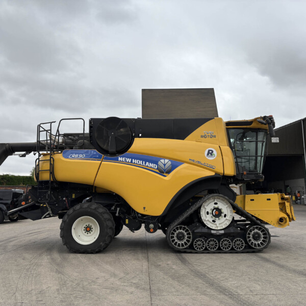 Yellow New Holland CR8.90 combine harvester with crawler tracks parked in an industrial yard against a cloudy sky, side view showing branding and machinery.