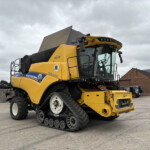 Yellow tracked combine harvester in a paved outdoor yard, cab visible with a brick building in the background.