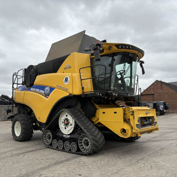 Yellow tracked combine harvester in a paved outdoor yard, cab visible with a brick building in the background.