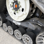 Close-up of a construction machine's rubber track and wheel assembly, showing a white wheel hub with orange center and bolts.