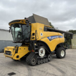 Yellow New Holland harvest machine on tracked wheels with a large front header, parked in an industrial yard under a cloudy sky.