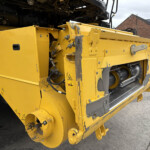 Close-up of a yellow road construction machine's front section with hydraulic components and warning labels, parked near a brick building.
