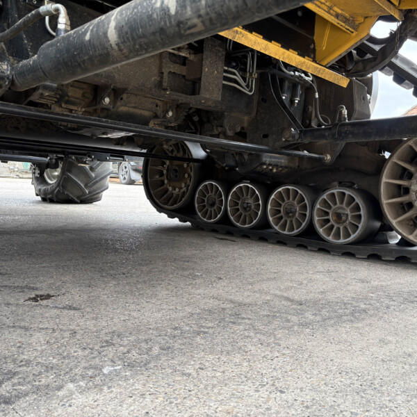 Underbelly of a muddy tracked vehicle, showing wheels, tracks, and exposed hydraulic lines and components under a yellow frame, low angle view.
