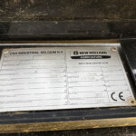 Close-up of a dusty metal data plate on a black surface; CNH Industrial Belgium N.V. and New Holland Agriculture branding visible.