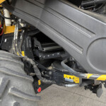 Close-up of heavy construction machinery showing hydraulic hoses, metal fittings, and a large black protective panel with a yellow frame nearby and warning labels visible.