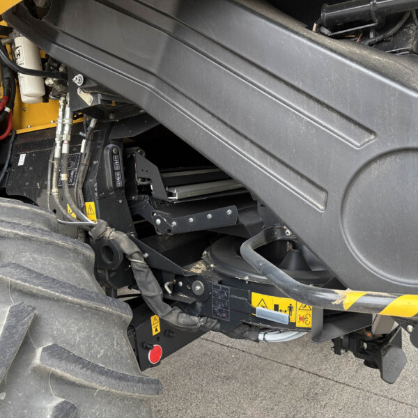 Close-up of heavy construction machinery showing hydraulic hoses, metal fittings, and a large black protective panel with a yellow frame nearby and warning labels visible.