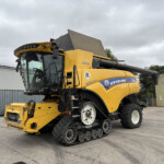 Yellow New Holland combine harvester on a paved lot, with a gray overcast sky and industrial buildings in the background.